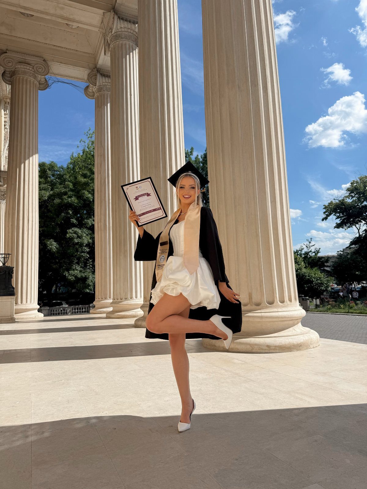 A graduate in a short white dress and black gown posing by classical columns.