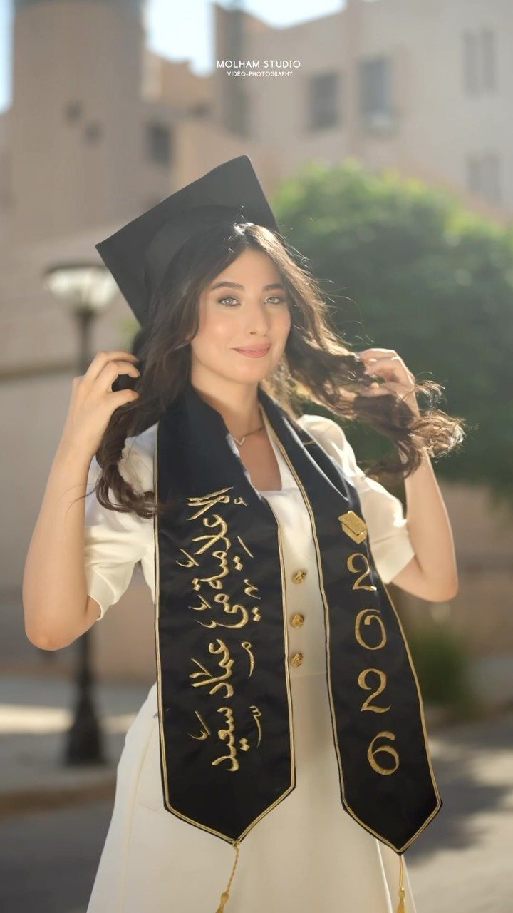 Smiling young woman wearing a white dress and a black graduation stole with gold lettering.
