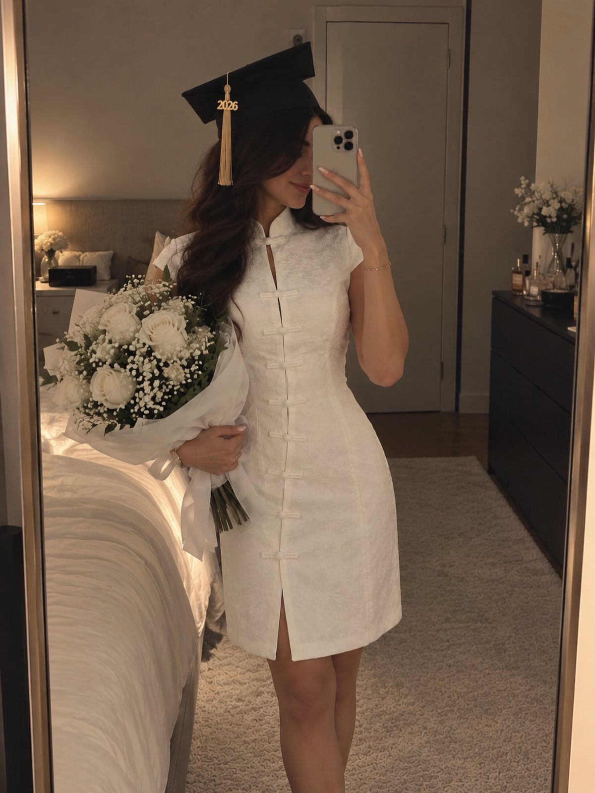 A woman in a white dress and graduation cap holding white flowers in a mirror selfie.